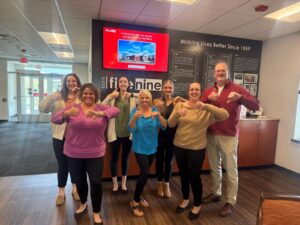 Eight coworkers in a bank lobby pose for a group photo, smiling and giving thumbs up.
